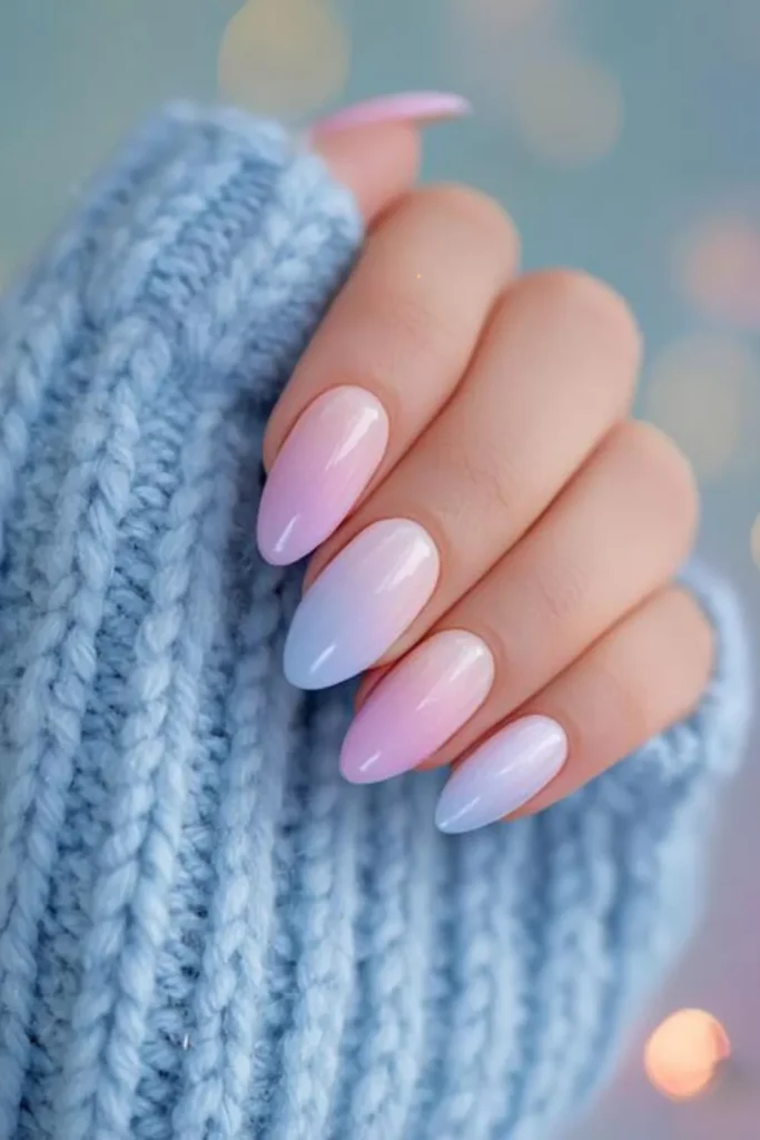 Glossy almond nails featuring a soft pastel gradient that transitions from light pink at the base to a pale blue at the tips, held against a blue knit sweater.