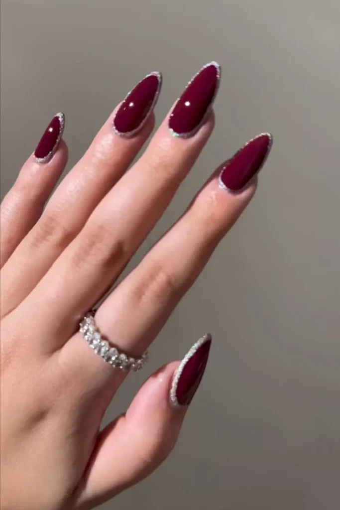 Long almond-shaped maroon nails with a delicate silver glitter border around the entire nail edge.