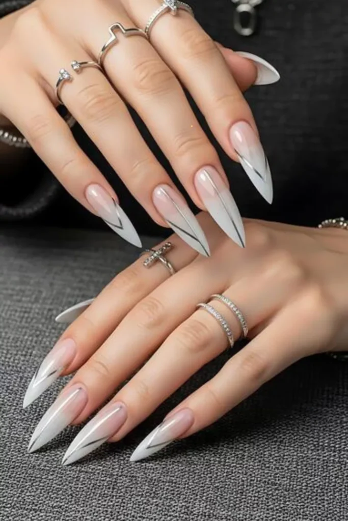Extra-long stiletto nails featuring a nude-to-white ombré base, decorated with sharp, minimalist silver metallic V-shaped lines and vertical stripes.