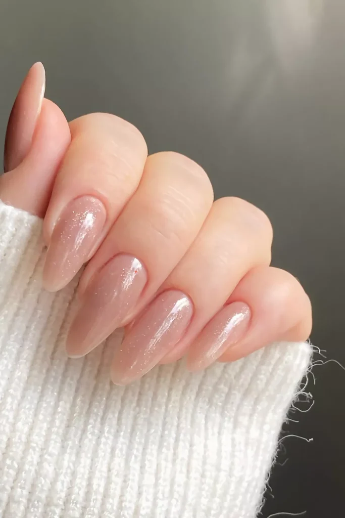 Long, almond-shaped nails featuring a high-shine, glossy nude polish with a fine, silver-toned shimmer overlay, held against a white textured knit sleeve.