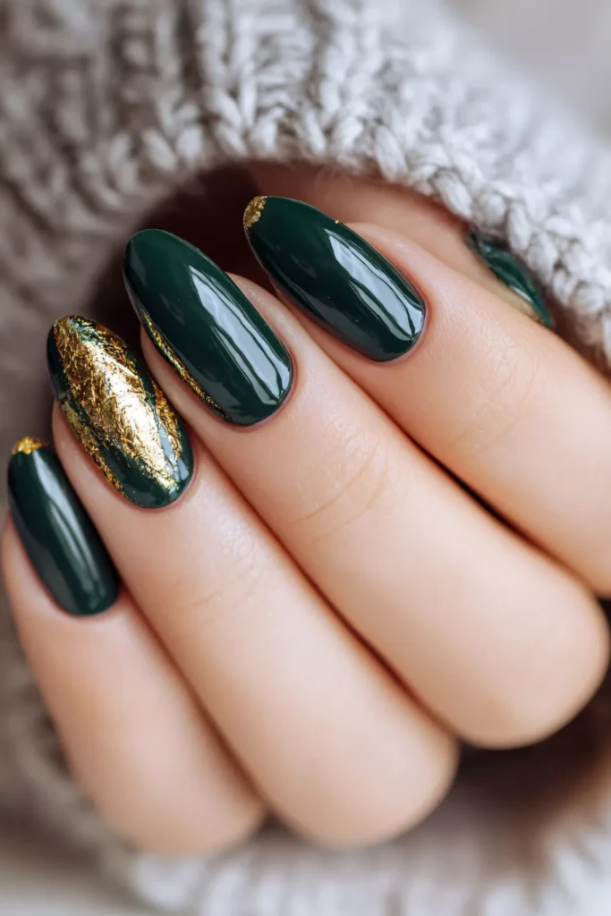 Close-up of glossy, deep forest green oval nails featuring luxurious gold leaf foil accents on the index and ring fingers.