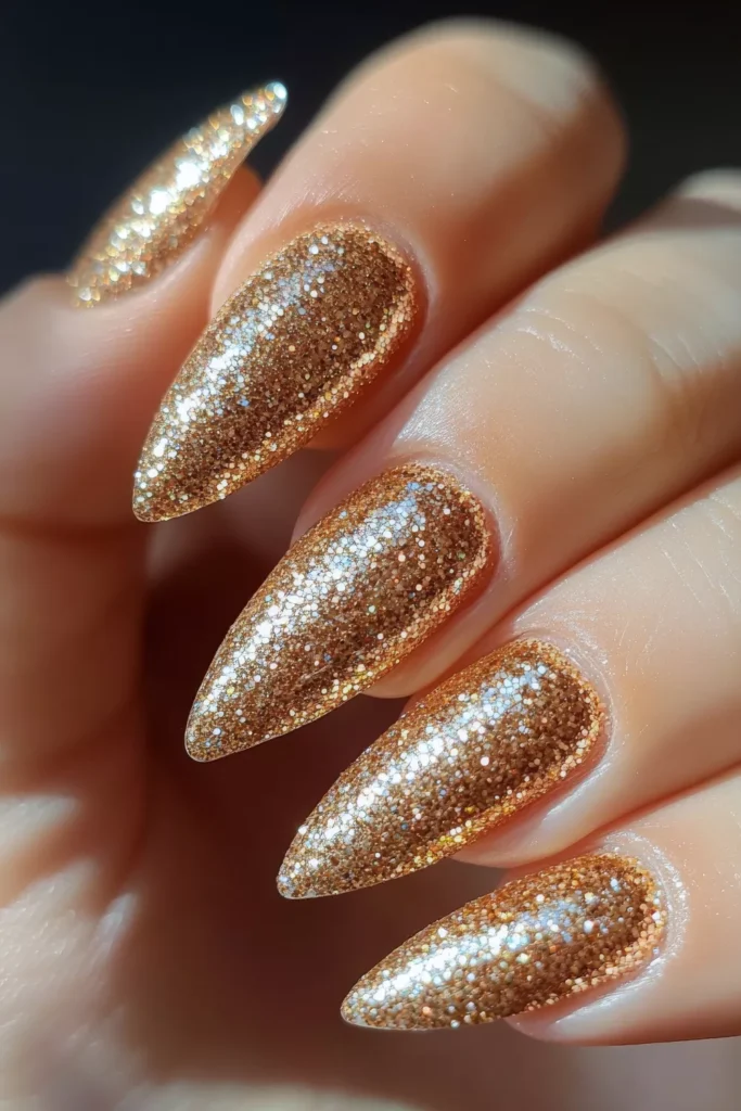 Long, stiletto-shaped nails completely covered in a dense, high-coverage bronze and gold micro-glitter polish that creates a textured, sparkling finish.