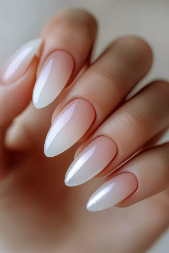 Long almond-shaped nails with a nude-to-white gradient ombre effect, featuring a pearlescent shimmer finish that catches the light on the tips.
