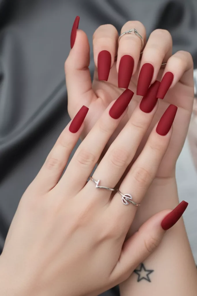 Long, matte red coffin-shaped nails styled with minimalist silver rings against a grey fabric background.