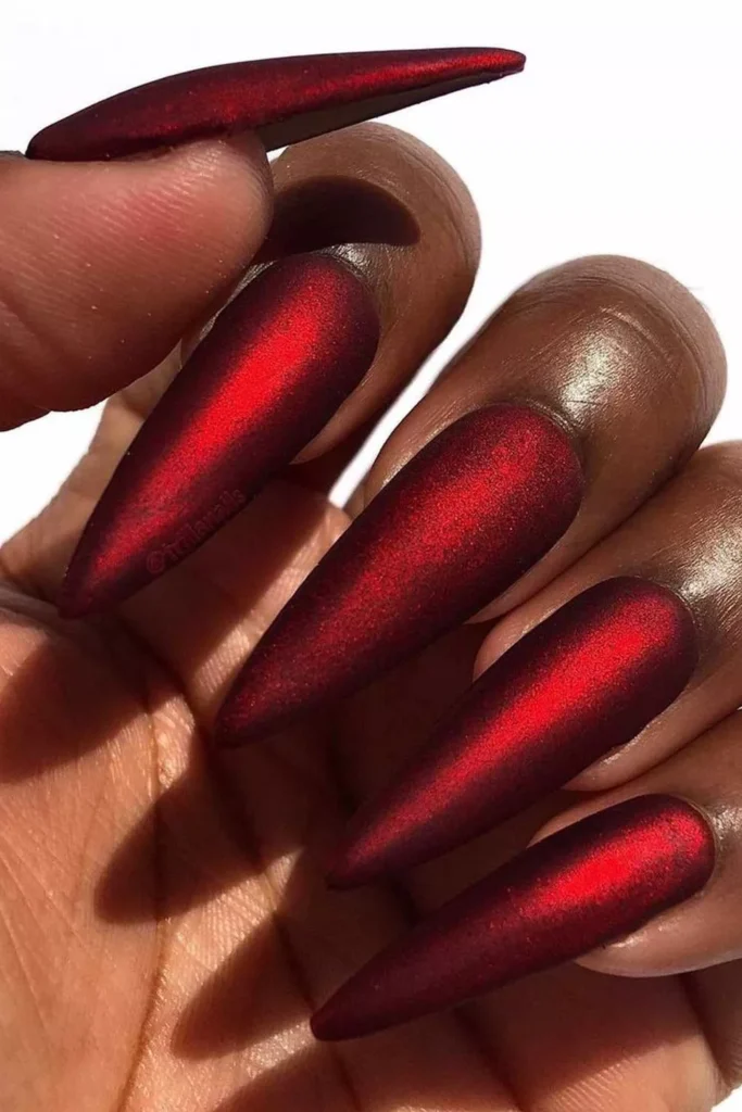 Long stiletto-shaped nails with a metallic deep red velvet finish on a dark skin tone.