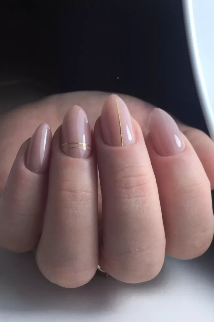 Glossy almond-shaped nails in a soft nude-pink shade, featuring minimalist gold metallic line accents positioned vertically and horizontally on the middle and ring fingers.