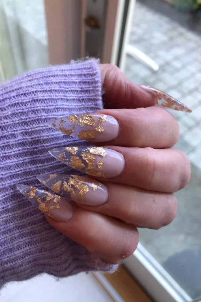 Long, pointed stiletto nails featuring a translucent nude base heavily decorated with textured gold leaf flakes, held against a purple knit sweater.
