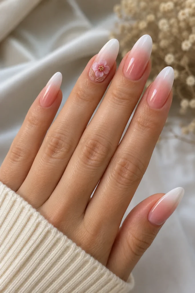 Almond nails with white-to-pink French ombre and a small 3D floral accent with gold detailing on the ring finger