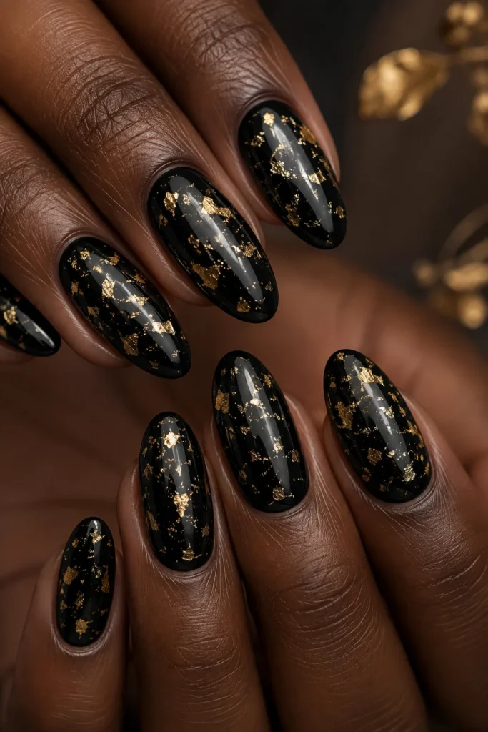 Glossy black almond nails covered in scattered gold foil flakes.