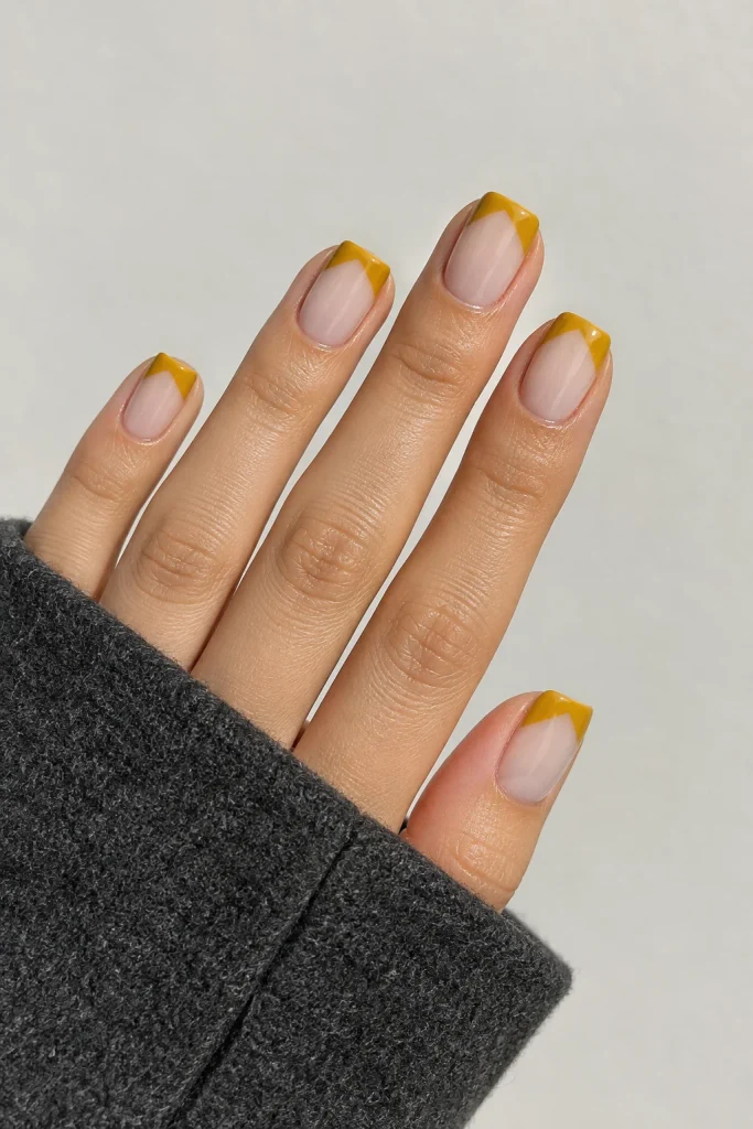 Short square nails with mustard yellow V-shaped French tips and nude base.